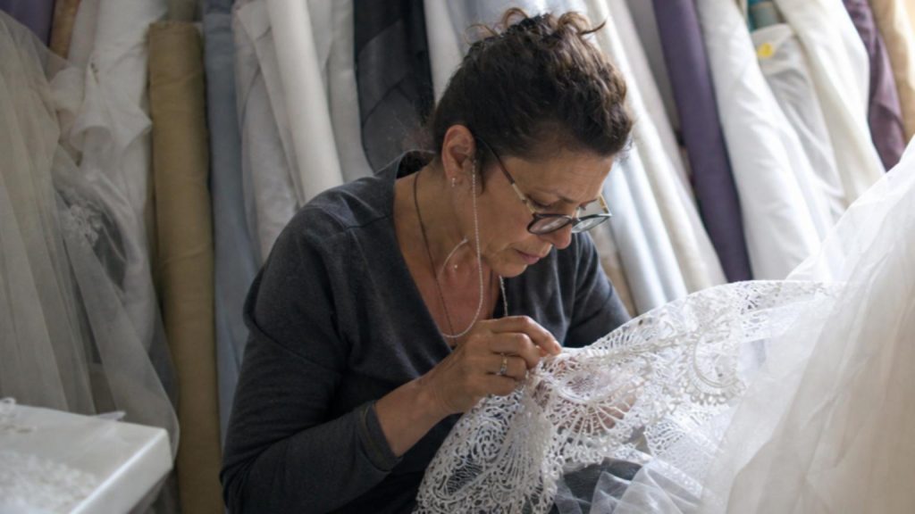 In a softly lit bridal atelier, a seamstress in the midst of a deli…