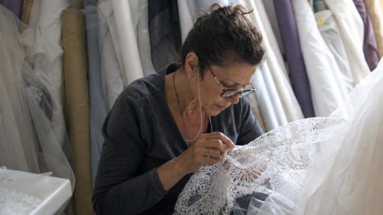 In a softly lit bridal atelier, a seamstress in the midst of a deli…